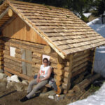 Photo: kleine Holzhütte mit neuem Dach im Schnee, dahinter Wald, vor der Hütte sitzt eine Frau mit Sonnenbrille in der Sonne