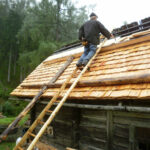Photo: Holzhütte auf einer Alm, Mann steht auf einer Leiter auf Höhe des Dachstuhls